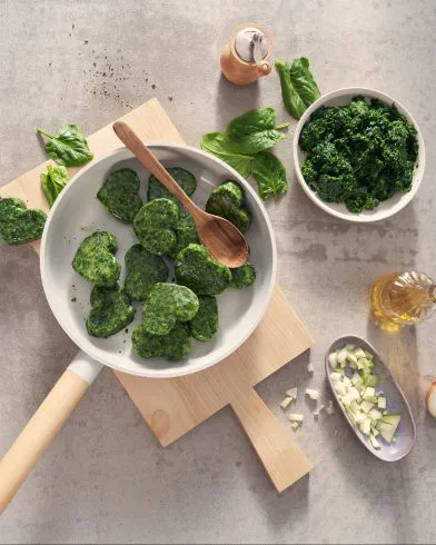 Na desce patelnia z zielonymi bryłkami mrożonego szpinaku FRoSTA, obok miska szpinaku, cebula, olej i młynek do pieprzu.
