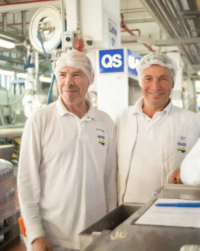 Dwóch mężczyzn w białych uniformach stoi wśród maszyn i produktów FRoSTA, uśmiechając się; na stole leży clipboard i długopis.