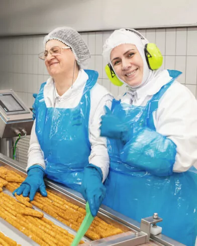 Dwie kobiety w odzieży ochronnej i fartuchach FRoSTA pracują przy linii, pakując mrożonki FRoSTA; śmieją się razem.