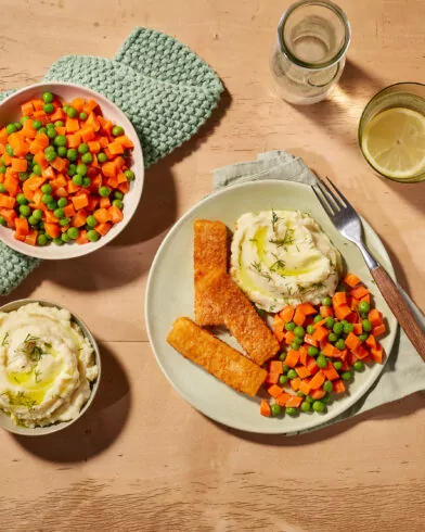 Talerz z puree i ziołami, paluszkami rybnymi FRoSTA, marchewką z groszkiem FRoSTA; obok miski z puree, warzywami FRoSTA i wodą z cytryną.