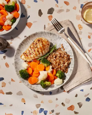 Talerz z grillowaną piersią kurczaka, kaszą gryczaną i bukietem warzyw FRoSTA na stole lastryko, obok sztućce i woda z cytryną.