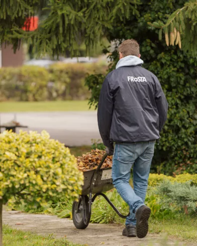 Osoba w ciemnej kurtce z logo FRoSTA wiezie taczkę z liśćmi i mrożonkami FRoSTA przez ogród w spokojnej okolicy.