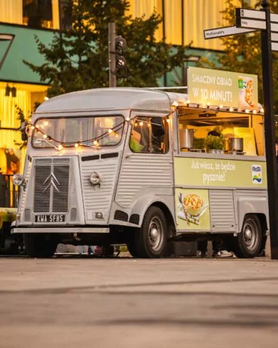 Szary food truck z lampkami oferuje dania i mrożonki FRoSTA, reklamowane na jasnym szyldzie po polsku, z kolorowymi zdjęciami.