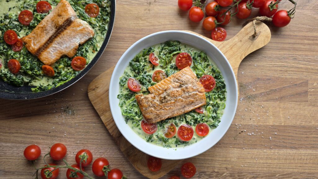 Miska z łososiem FRoSTA w sosie szpinakowym na kremowym szpinaku i pomidorach, obok patelnia z daniem FRoSTA.