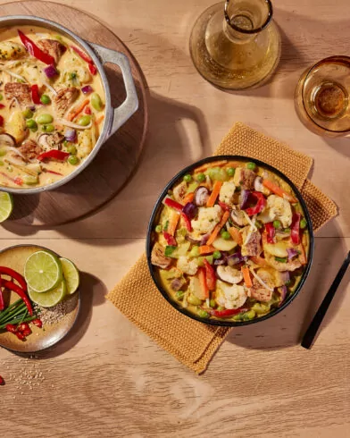 Garnek i miska kremowego curry z warzywami FRoSTA, tofu, groszkiem, papryką i cebulą na stole obok napoju, limonek i przypraw.