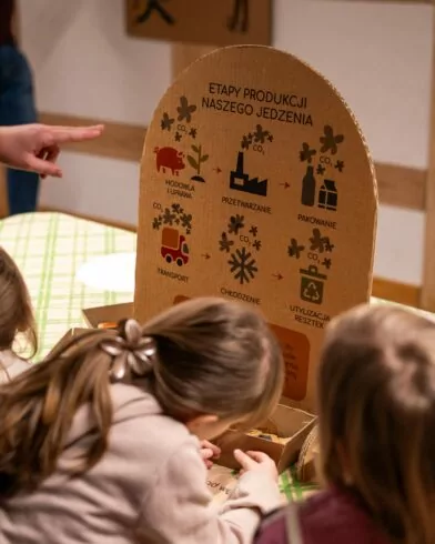 Troje dzieci ogląda planszę o etapach produkcji z mrożonkami FRoSTA, a dorosły wskazuje ilustracje i polskie napisy na stole.