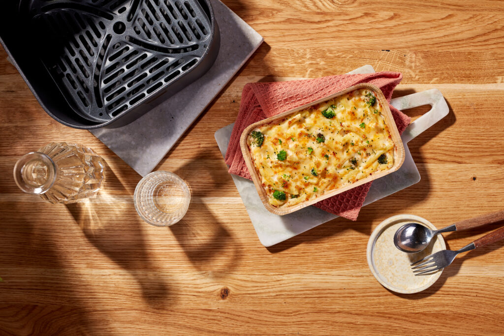 Zapiekanka makaronowa z brokułami, obok kosza do frytkownicy FRoSTA AIR FRYER oraz mrożonego produktu FRoSTA na desce, dzbanek, kubek i talerzyk ze sztućcami.